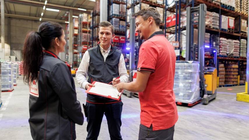 Logistik-Mitarbeiter von Kaufland unterhält sich im Lager mit seiner Kollegin und seinem Kollegen und macht sich Notizen auf seinem Clip-Board.